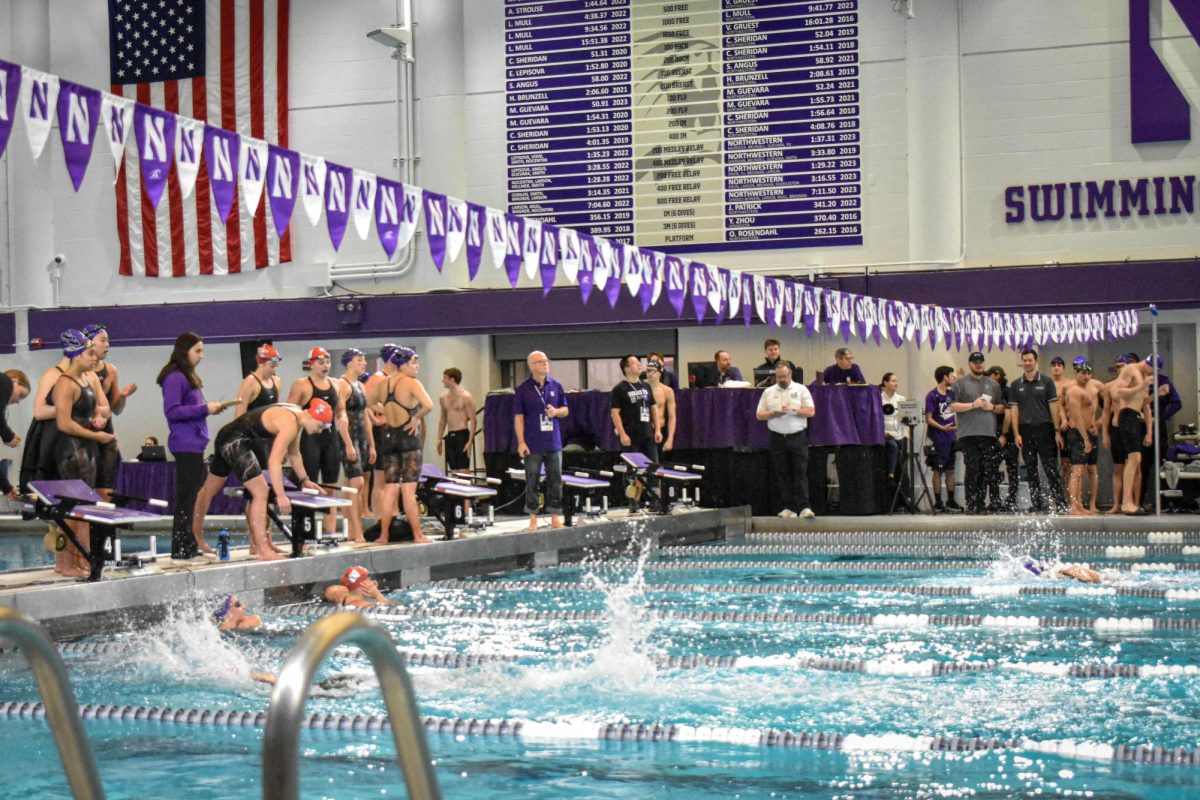 Wisconsin and Northwestern women’s swim teams race against each other, as teammates from both teams observe on the sidelines, some preparing to swim next. Judges in purple stand on the sidelines and sit behind a purple-clothed table.