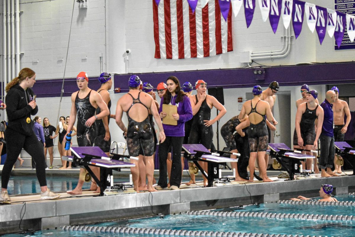 A Northwestern swim coach in a purple jacket confers with a female Northwestern swimmer donning a swim cap, goggles, and a suit.