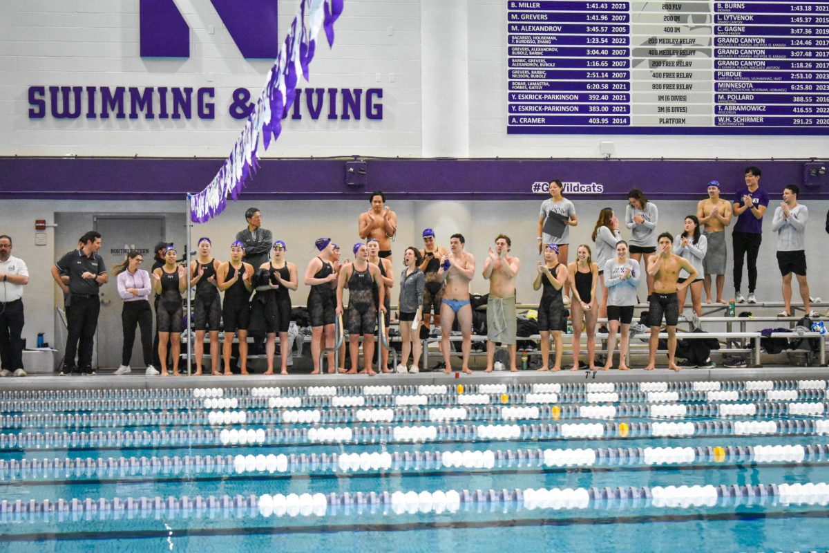 Teammates of Northwestern Men’s and Women’s teams clap and cheer on their teammates while standing in their swimsuits in the bleachers.