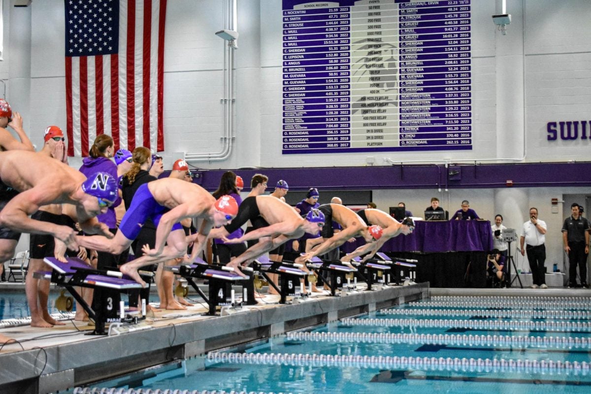 Two Northwestern male swimmers get in position on the racing platform to race two Wisconsin male swimmers, with a fifth unidentified swimmer in the back. Judges in purple stand on the sidelines and sit behind a purple-clothed table.