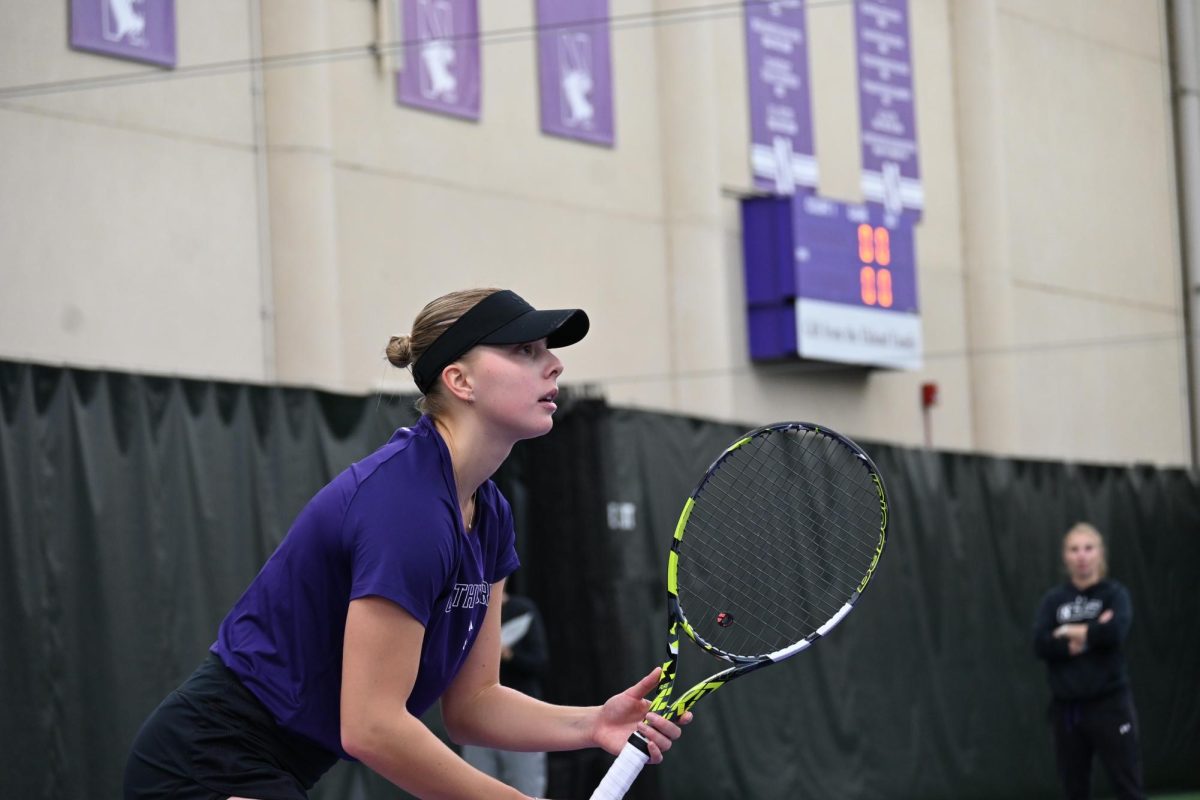 An NU Women’s Tennis player waiting for the ball as a person observes in the background.