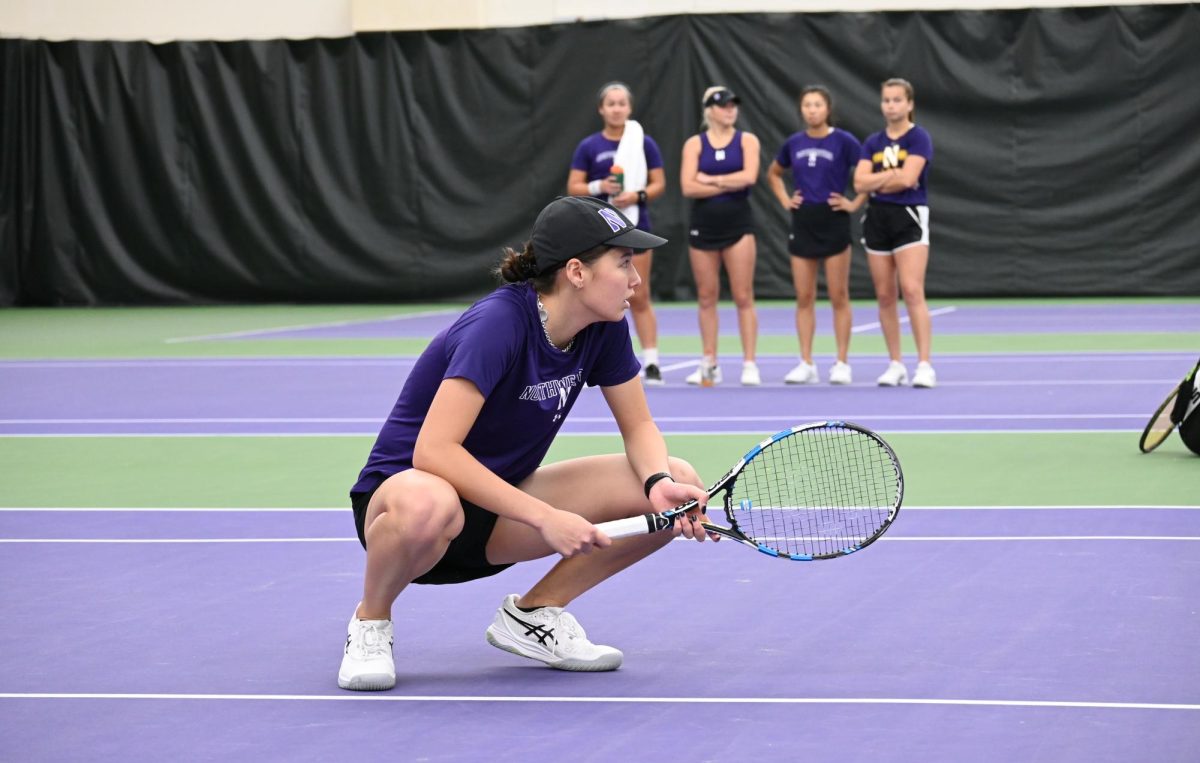 An NU Women’s Tennis player waiting for her doubles partner to serve as four NU tennis players observe.