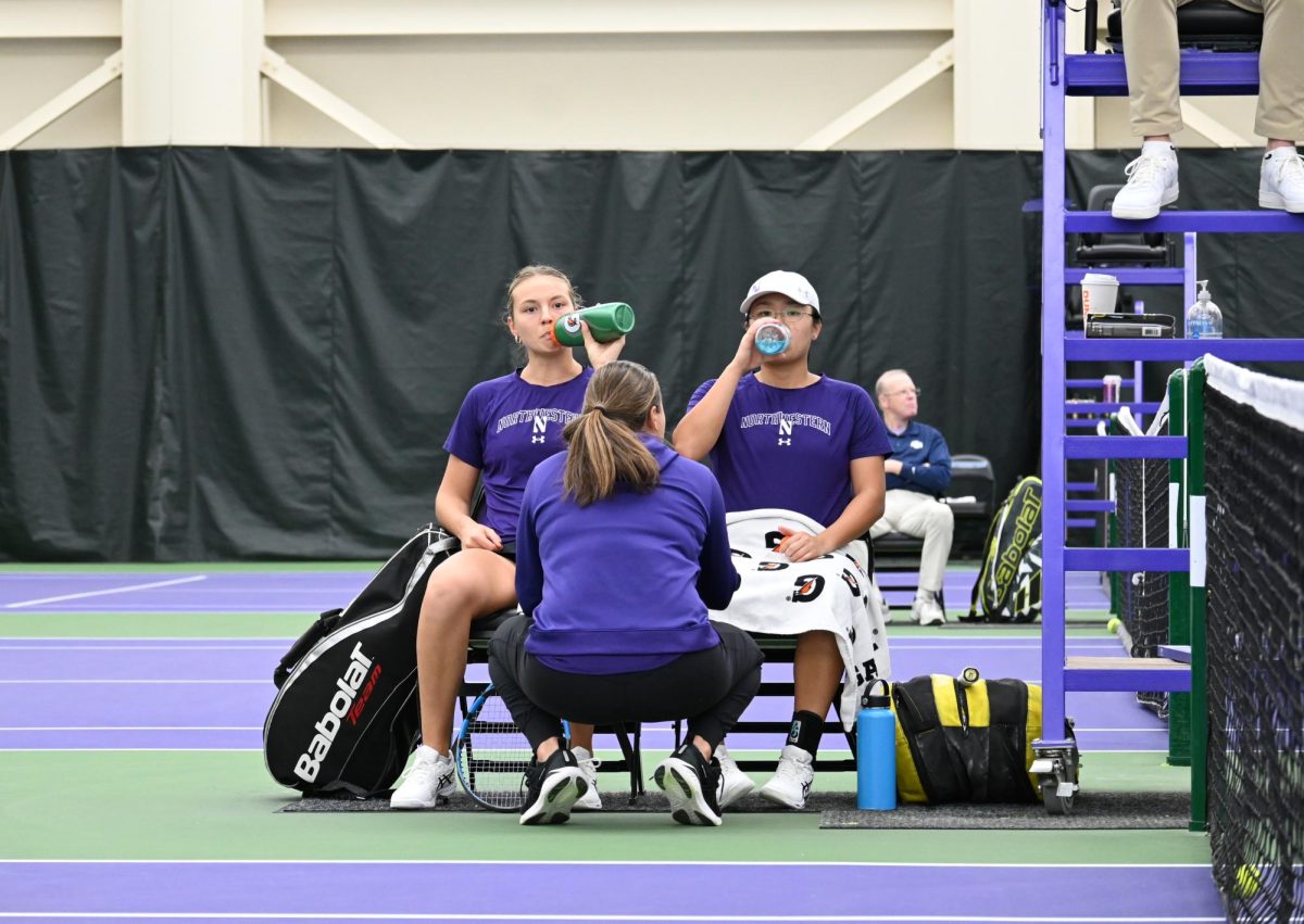 wo NU Women’s Tennis players and a coach talking over a break between sets while sipping from their water bottles.