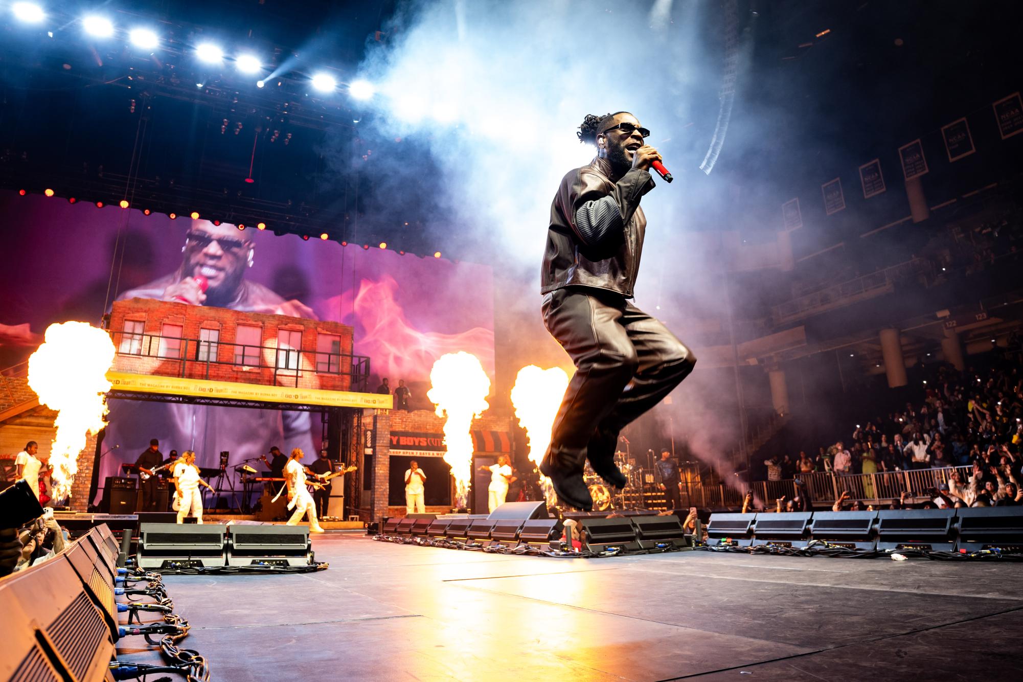 A singer wearing all black and sunglasses jumps as flames shoot up from the stage behind them.