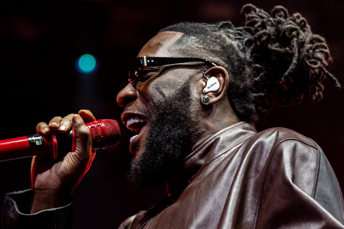 A singer wearing sunglasses with an earplug with ‘Burna’ on it performs with a red microphone.