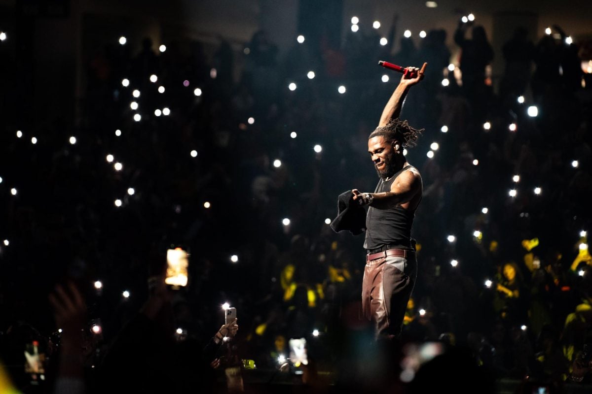 A singer wearing a black shirt and brown pants raises the red microphone as cellphone flashlights shine in the background.