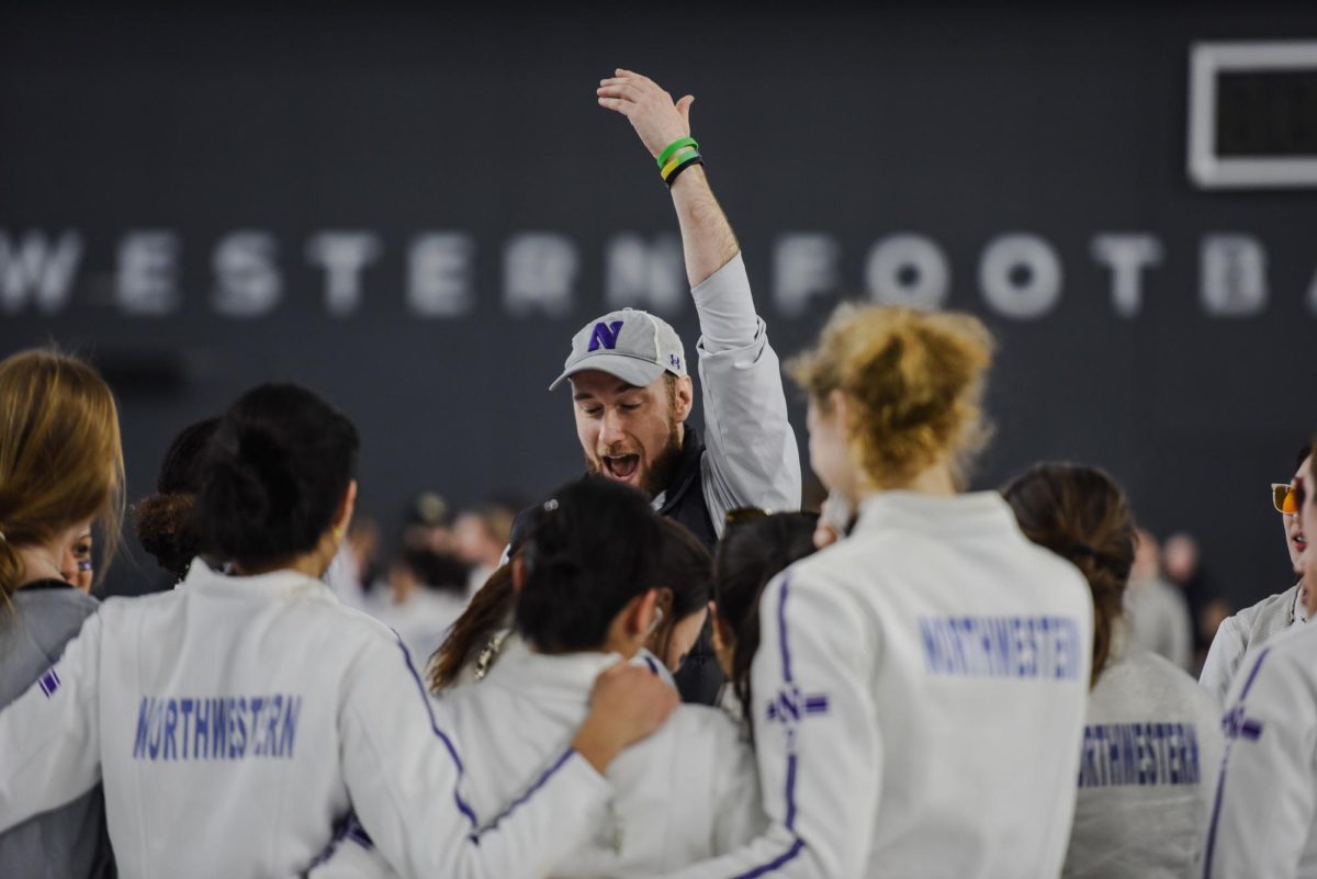 Northwestern fencing coach Zach Moss wearing a gray hat holds his hand in the air while surrounded by athletes in purple and white uniforms.