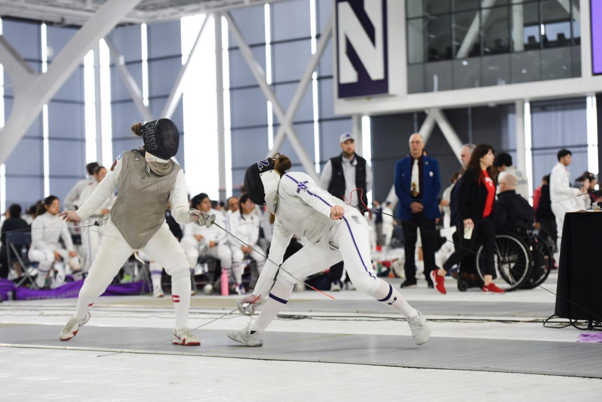 A Northwestern fencer in white and a black mask holds a saber and swipes at their opponent’s leg with a saber while their opponent in gray strikes them with a saber.