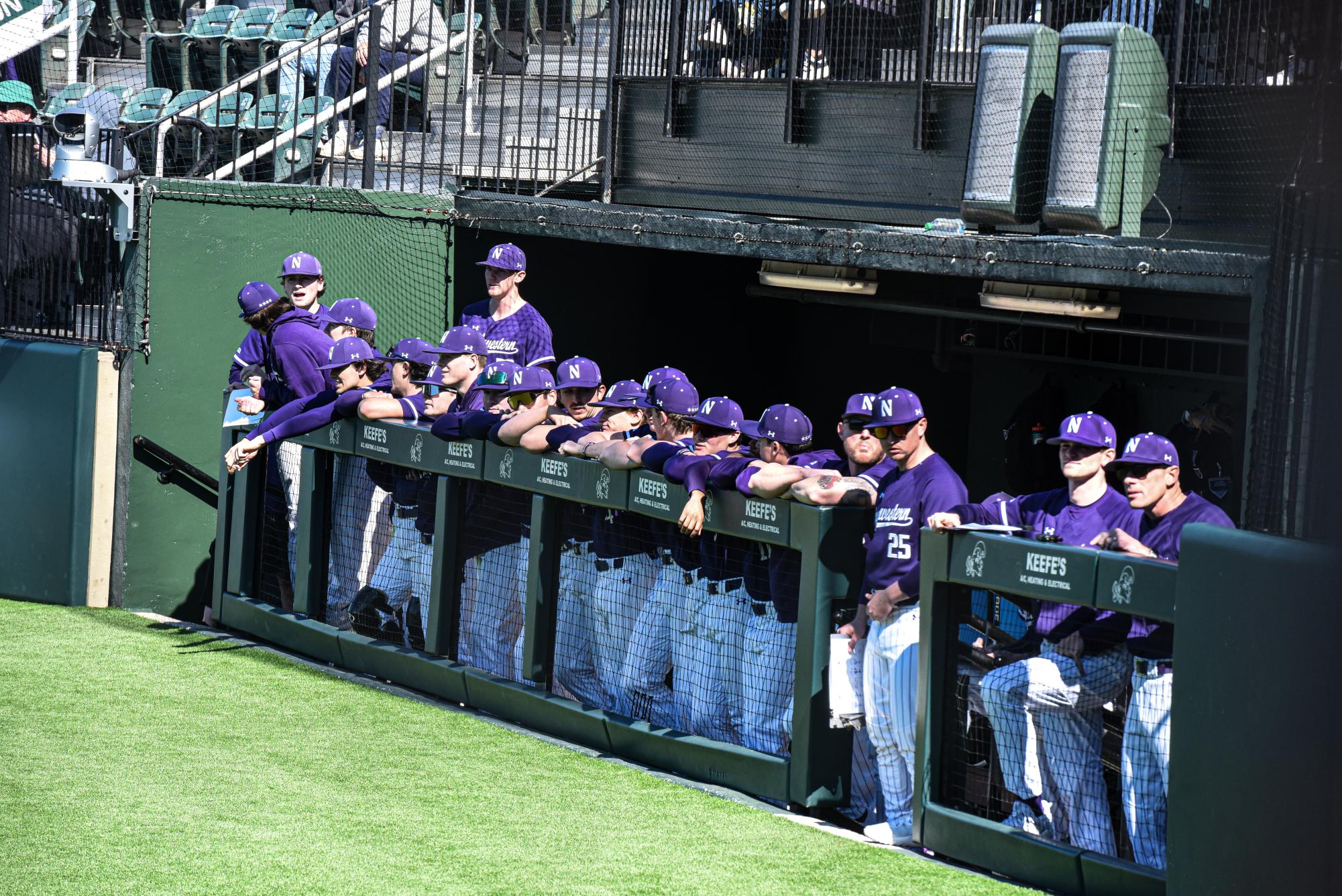 Alt Text: Northwestern baseball players wearing purple stand in the dugout.