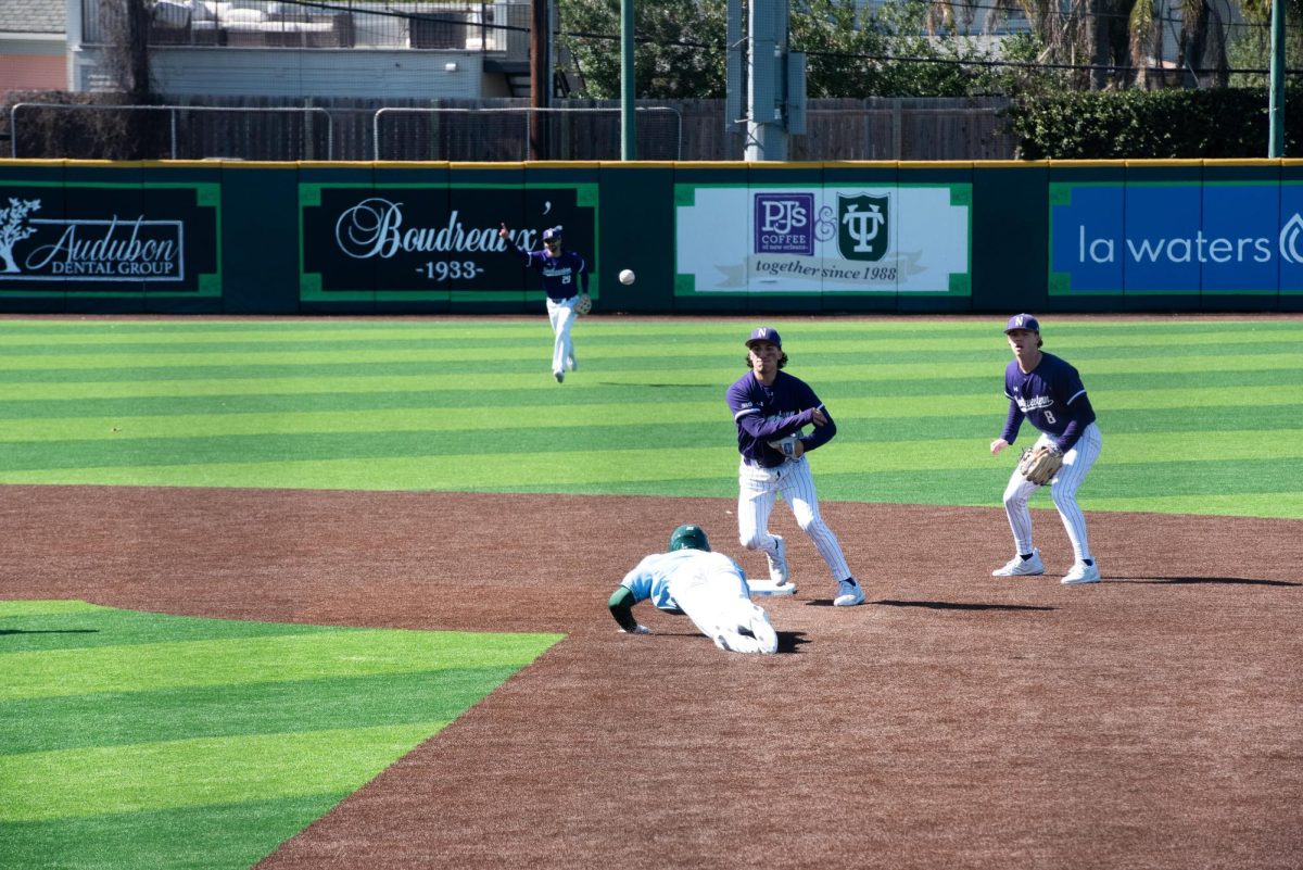 A Northwestern baseball player, wearing purple, throws a baseball while a Tulane baseball player, wearing blue and green, slides to second base. Players also stand in the outfield. 