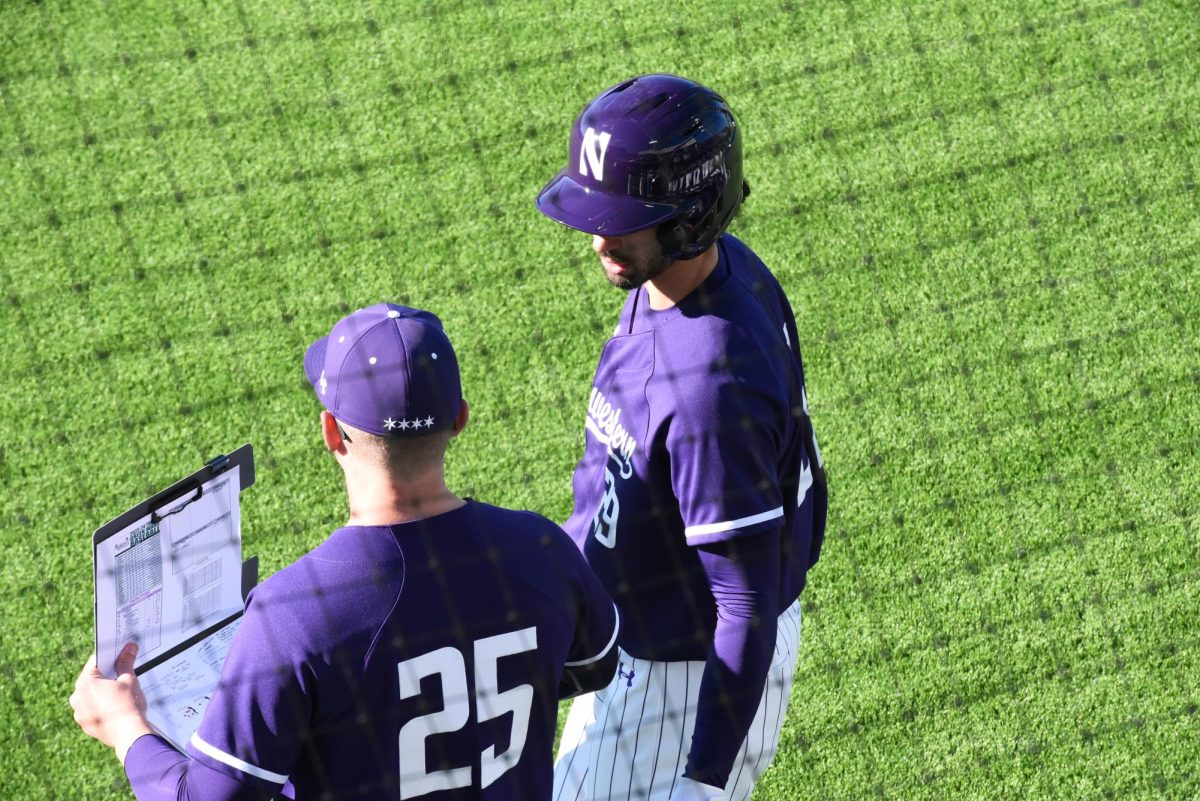 A Northwestern baseball player wearing purple talks to a Northwestern coach who is wearing a purple 25 jersey and holding a clipboard. 