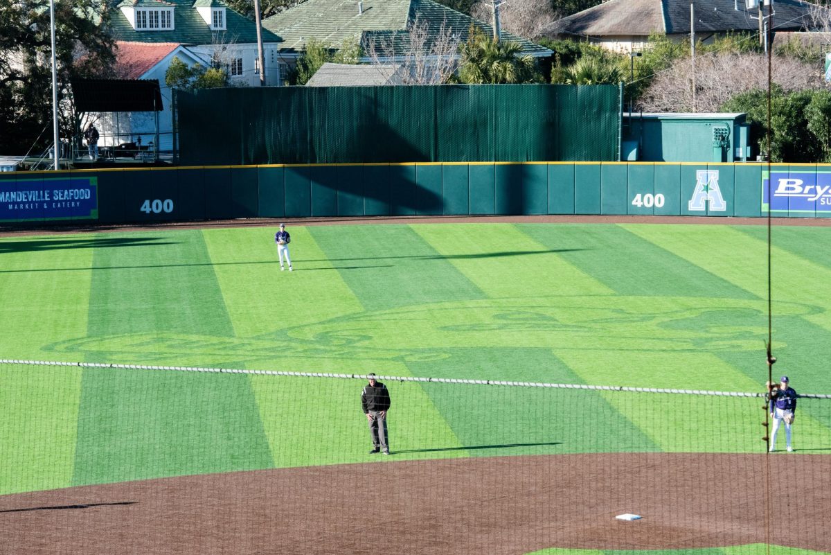 A Northwestern baseball player wearing purple stands in center field next to Tulane’s logo, while an umpire and a Northwestern baseball player stand infield.