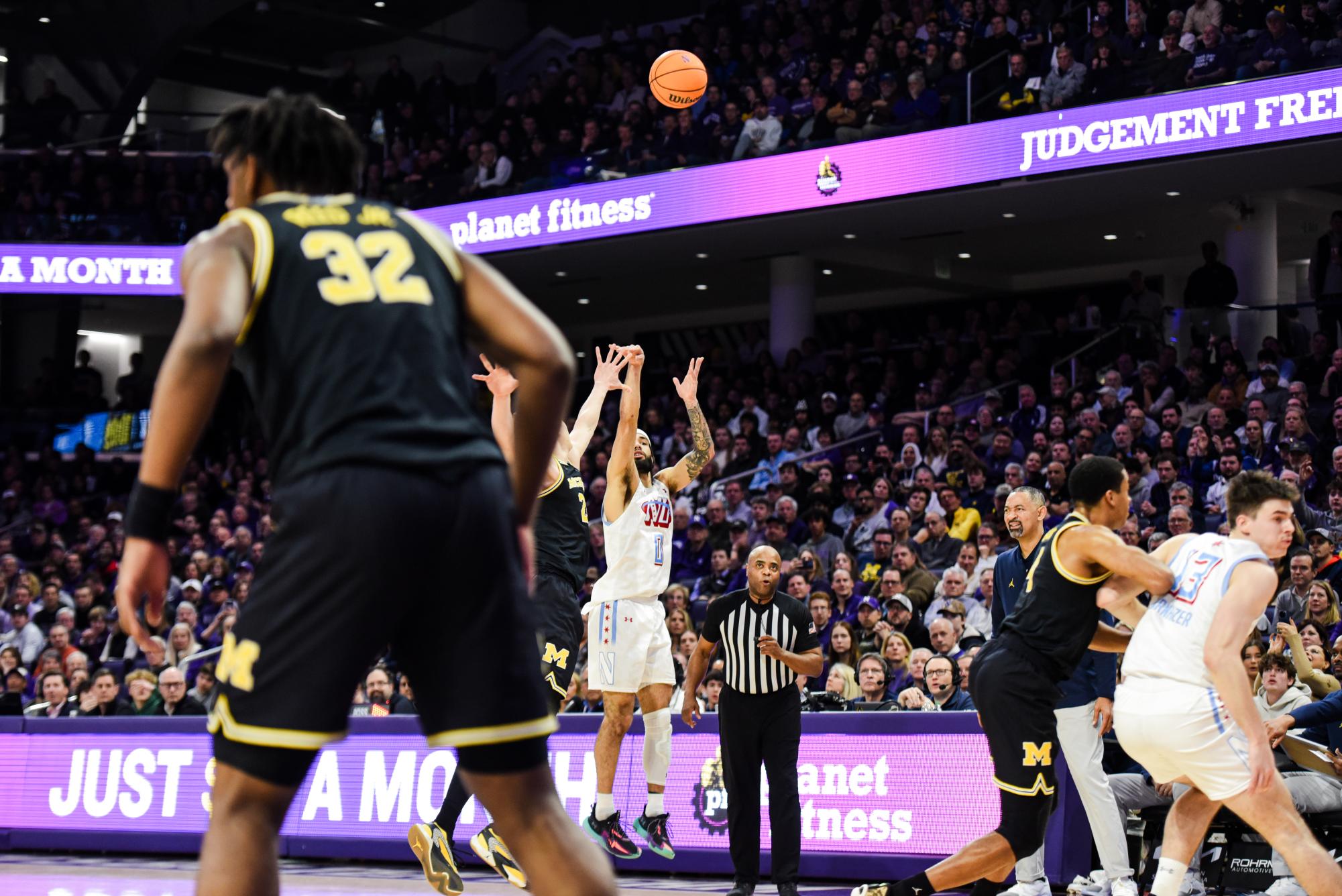Northwestern’s Boo Buie wearing white shoots a three pointer while being defended by a basketball player wearing black and yellow.