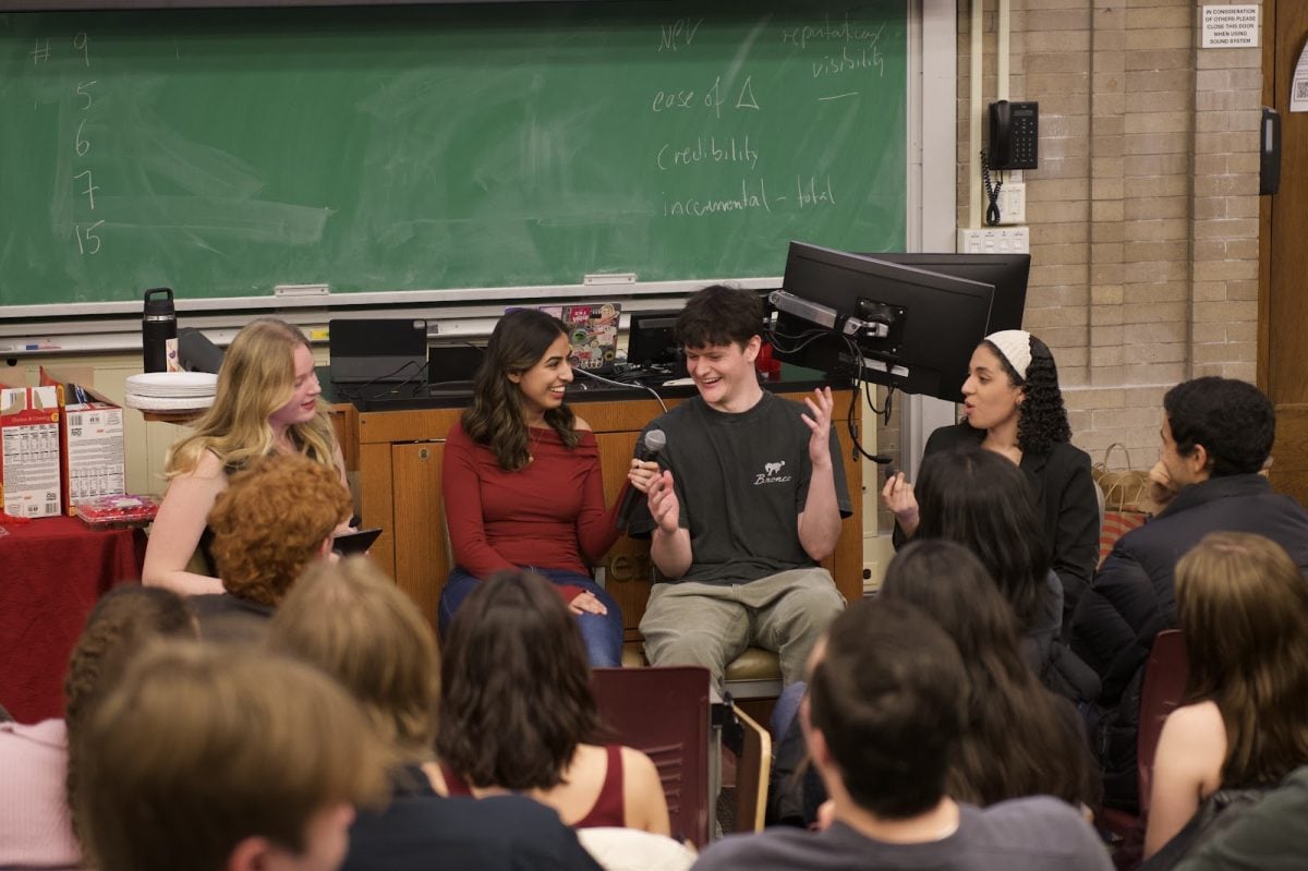 Four people sit and talk at the front of a crowded lecture hall, and one person holds a microphone.