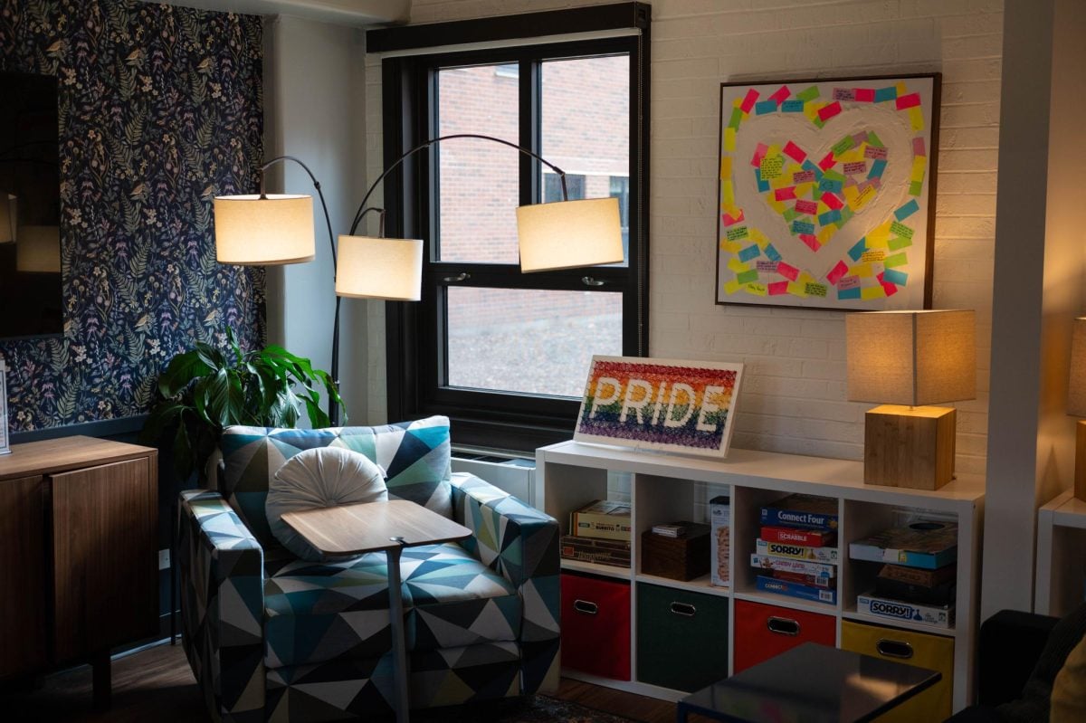 An armchair stands by the window next to a bookcase filled with board games. A colorful sign that reads “pride” in all-uppercase letters sits on top of the bookcase next to a wooden lamp.