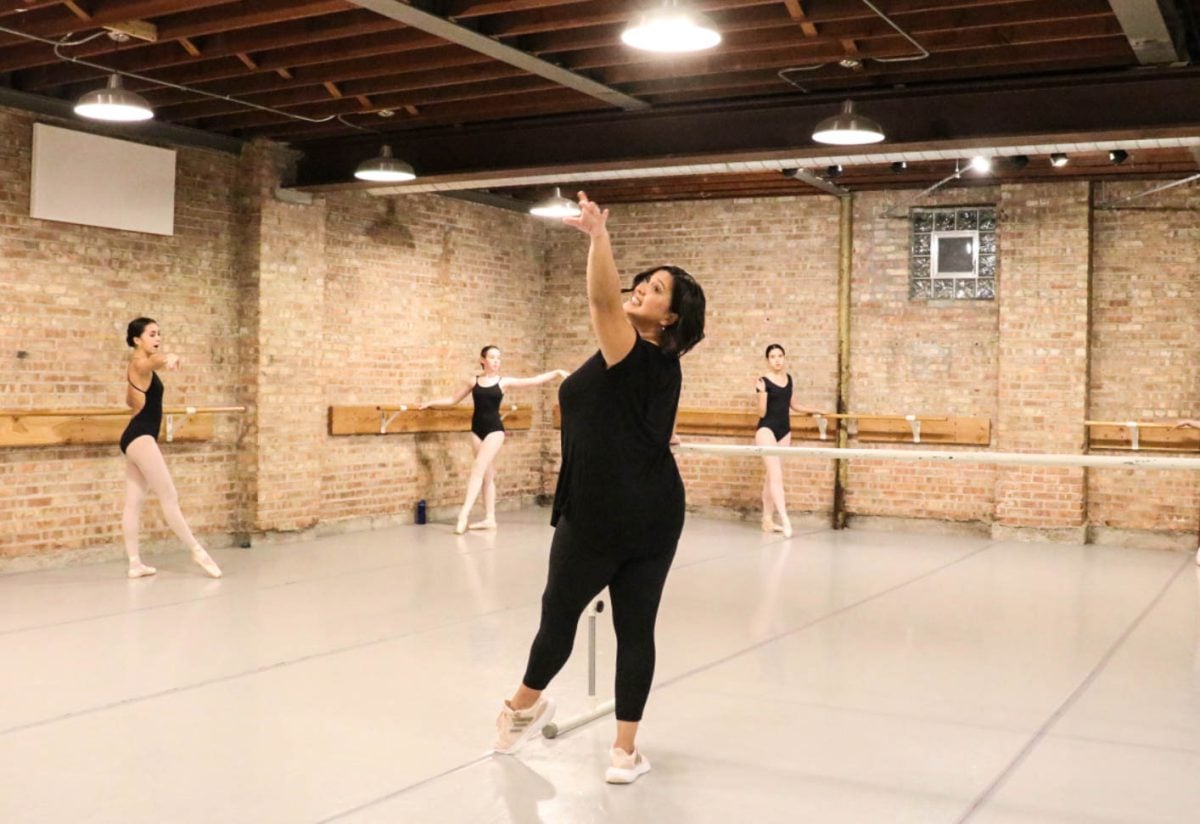 Maliwan Diemer stands in a front tendu in the center of a brick dance studio at a barre, leading her students in the background of the image in a barre routine.
