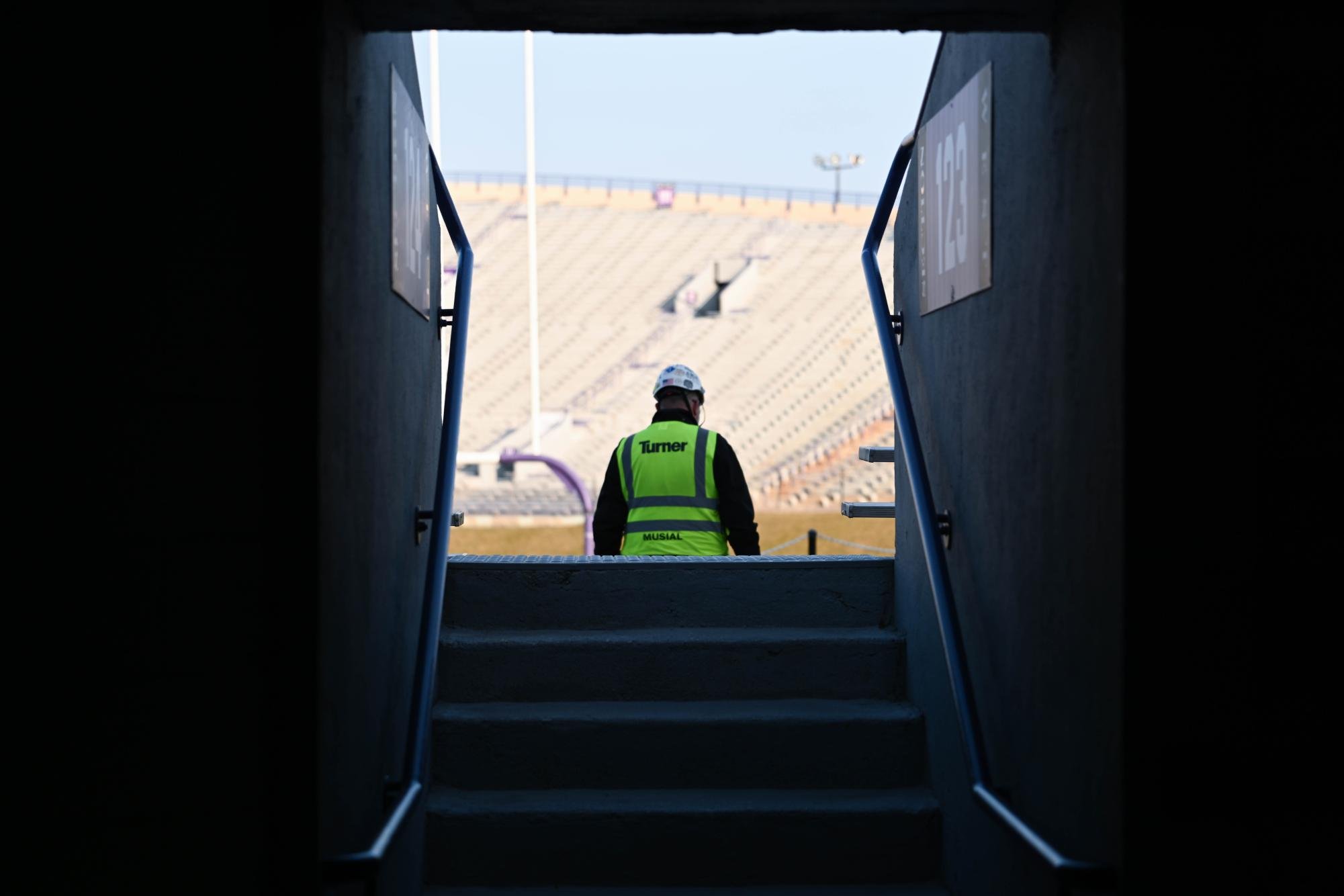 A man wearing a hard hat looks out at bleachers in a football stadium.