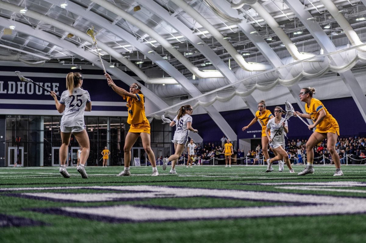 Taylor with the ball as a Marquette defender approaches her, holding her lacrosse stick in the air. There are other Northwestern attackers and Golden Eagle defenders to her left. 