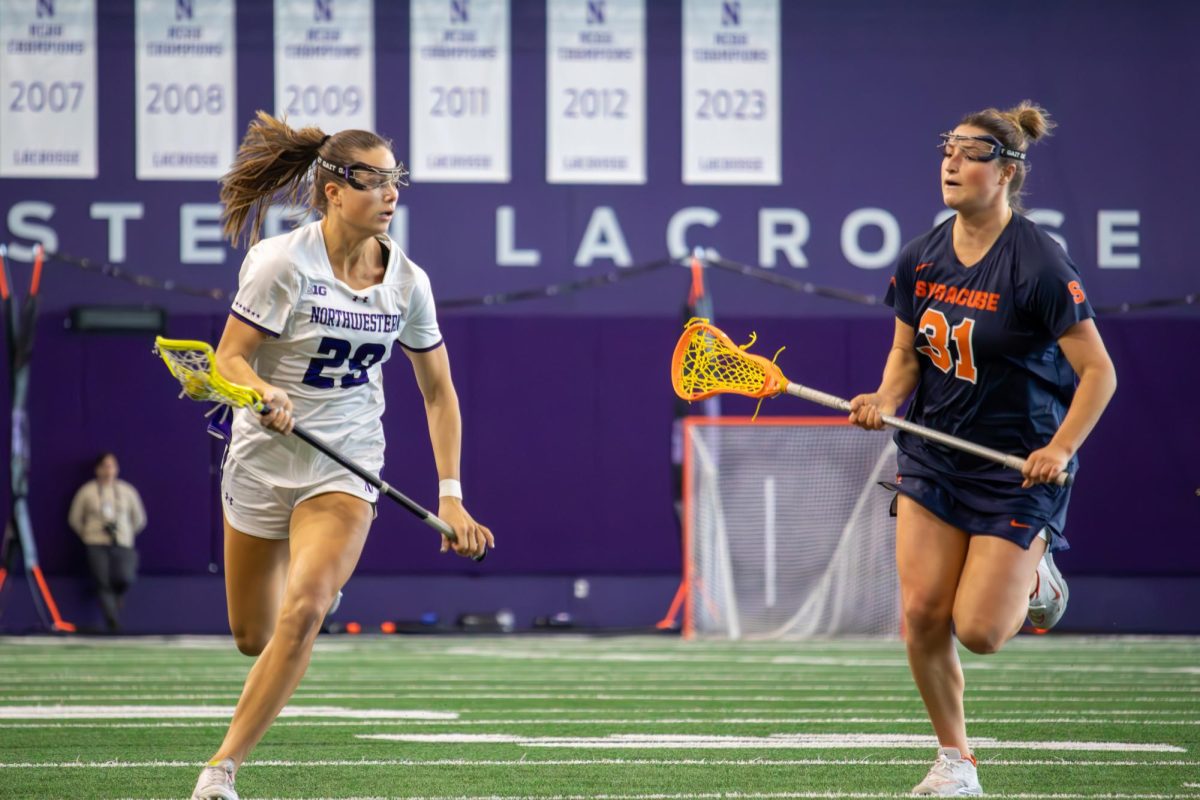 Senior defender Carleigh Mahoney runs forward with a lacrosse stick and ball as an opponent in a blue and orange uniform runs to the side.