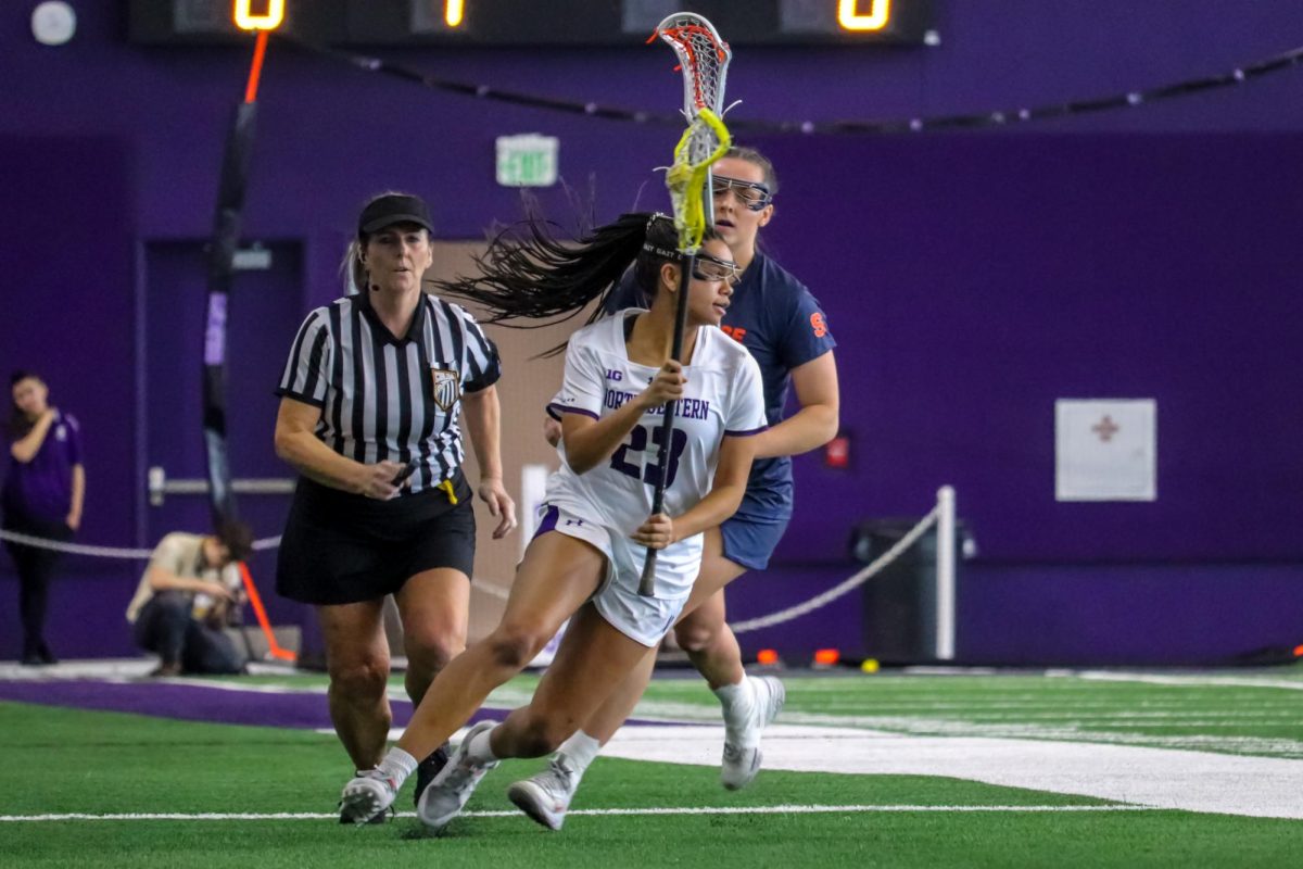 Junior defender Sammy White, wearing a white and purple uniform, runs with a lacrosse stick and ball as a referee and opponent wearing a blue and orange uniform trails behind.