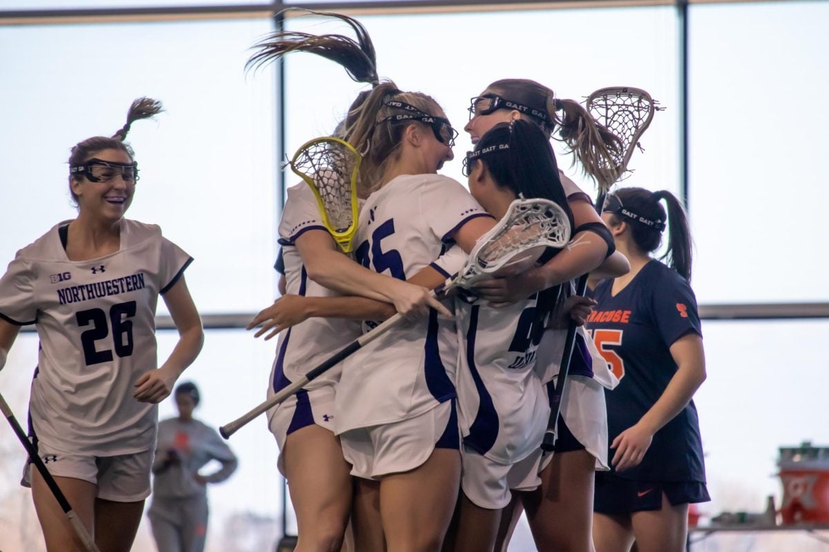 Five Northwestern lacrosse players hug and celebrate while holding their lacrosse sticks.