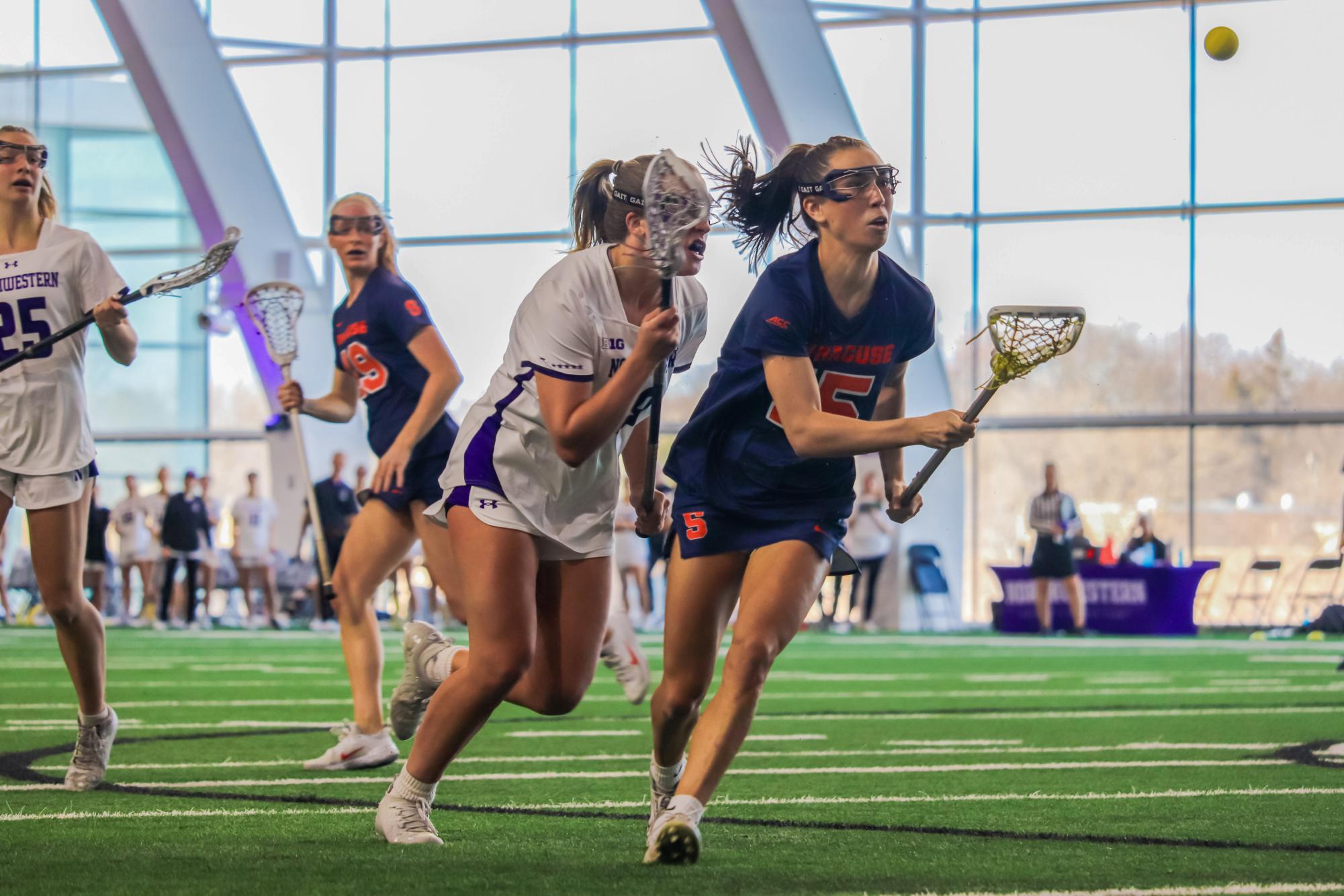 A lacrosse player in a white and purple uniform and another in a blue and orange uniform run with lacrosse sticks as they chase a yellow ball in the air.