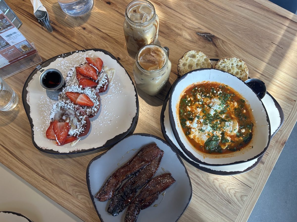 A wooden table with plates of shakshuka, bacon, and red velvet french toast, along with two mason jar glasses of iced coffee.