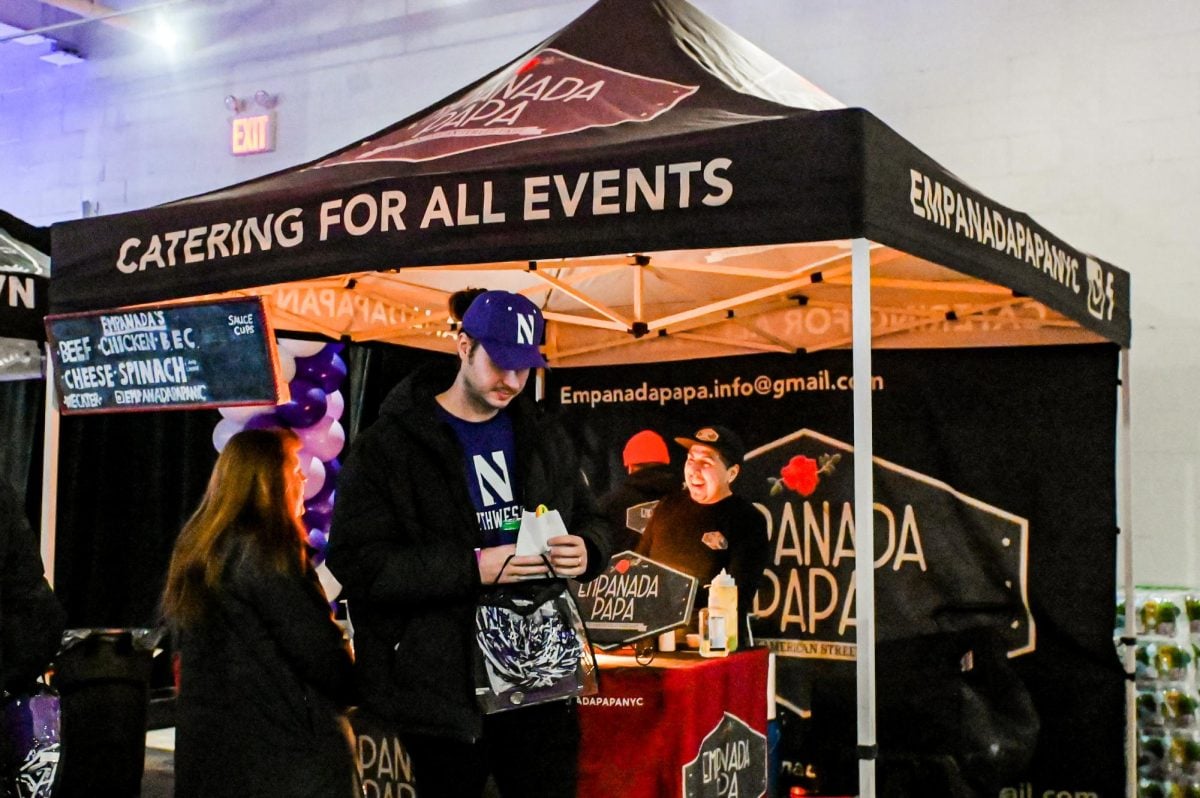 A person eats an empanada. A person in the background in a black tent greets the next customer in line.