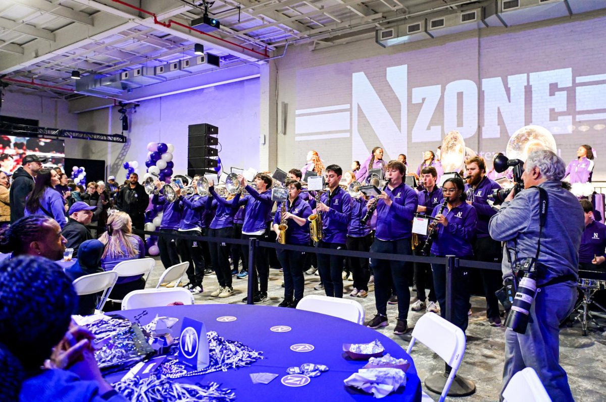A marching band in purple plays while cheerleaders behind them on a stage dance. The phrase “N zone”is shown in the back.