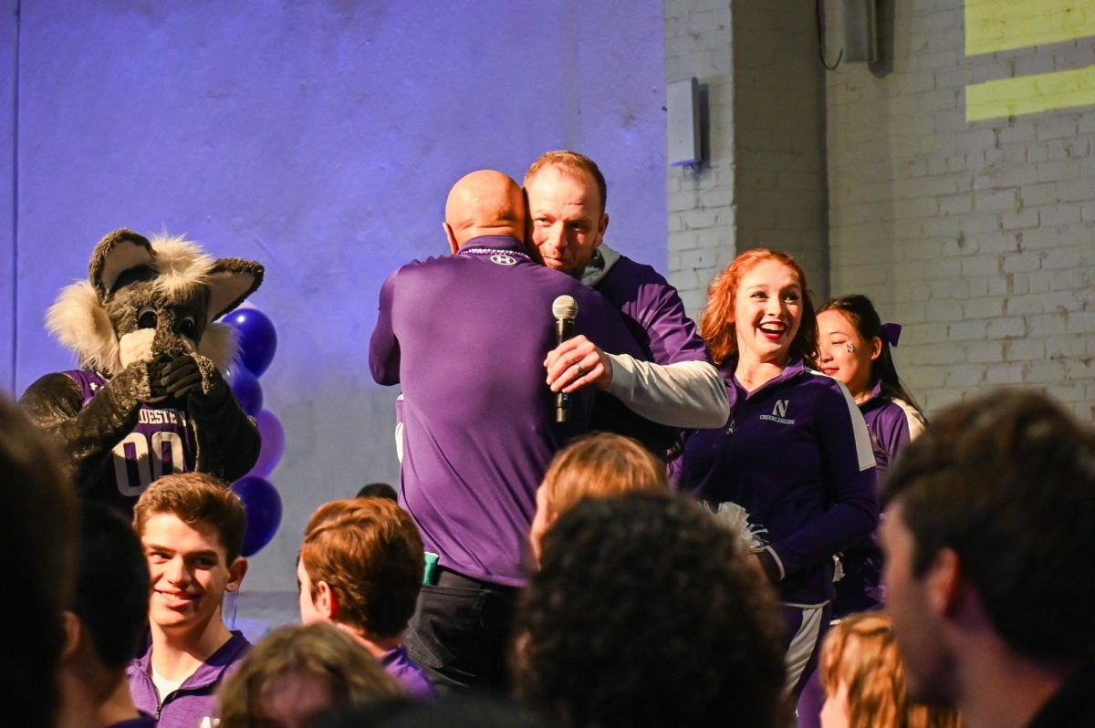 Two people in purple embrace on a stage while others around them cheer.