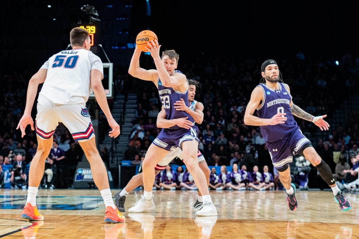A basketball player in purple attempts to throw the ball to another player while an opponent player in white wraps their arms around them in a foul.