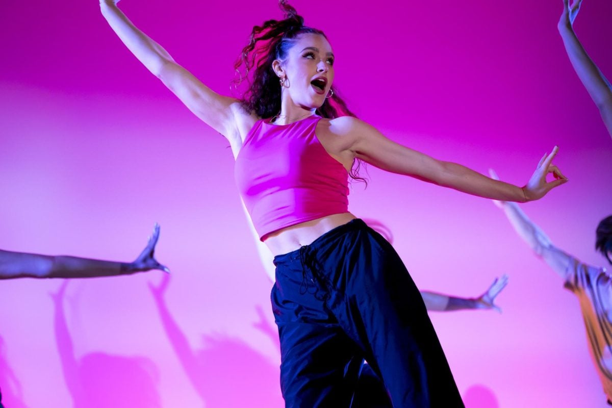 A dancer in a pink crop top and black joggers dances with arms outstretched in front of a pink-lit background.