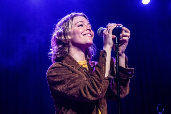 Maggie Rogers, wearing a leather jacket and a yellow shirt, smiles in front of a microphone.