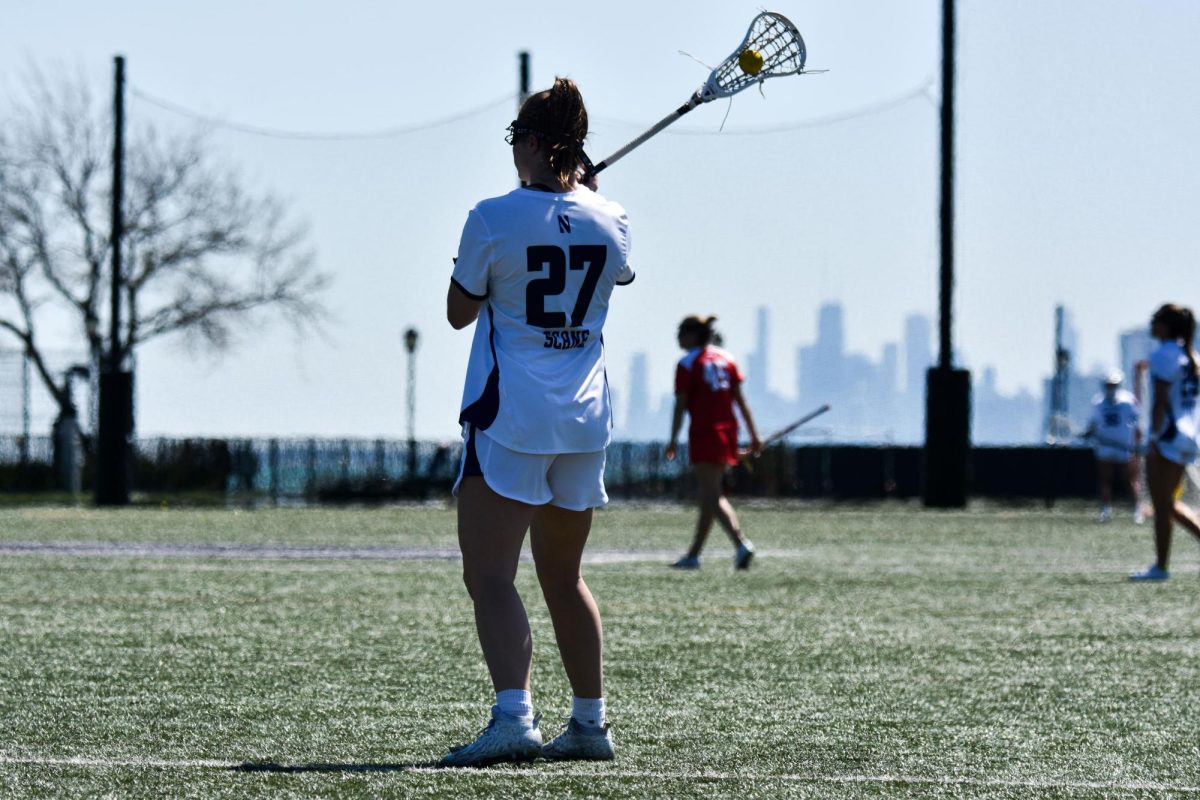 A Northwestern lacrosse player wearing white holds the lacrosse ball in her stick and looks toward the field.