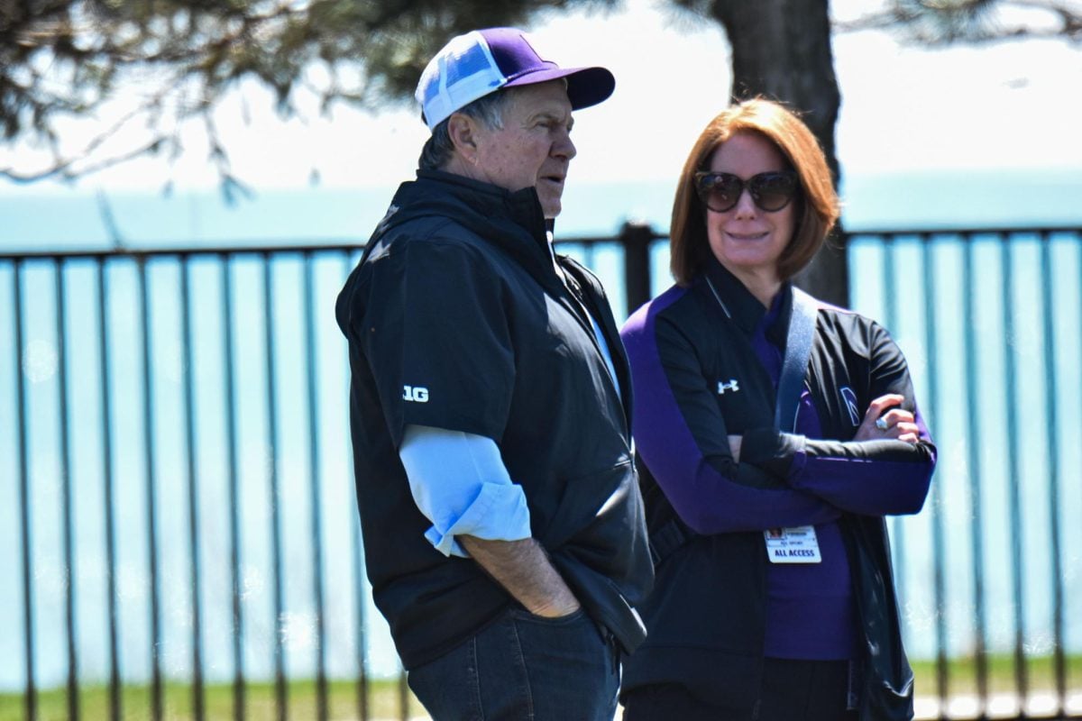 Bill Belichick, wearing a black shirt and a purple hat, watches lacrosse with a person wearing black and purple to his side.