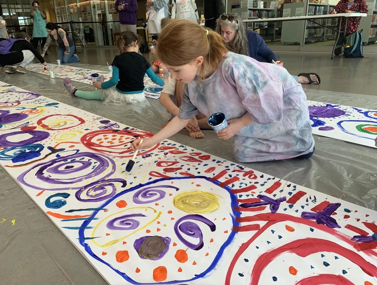 A child in a tie-dye smock kneels on the ground, painting blue dots on a paper filled with bright painted dots, circles and spirals.