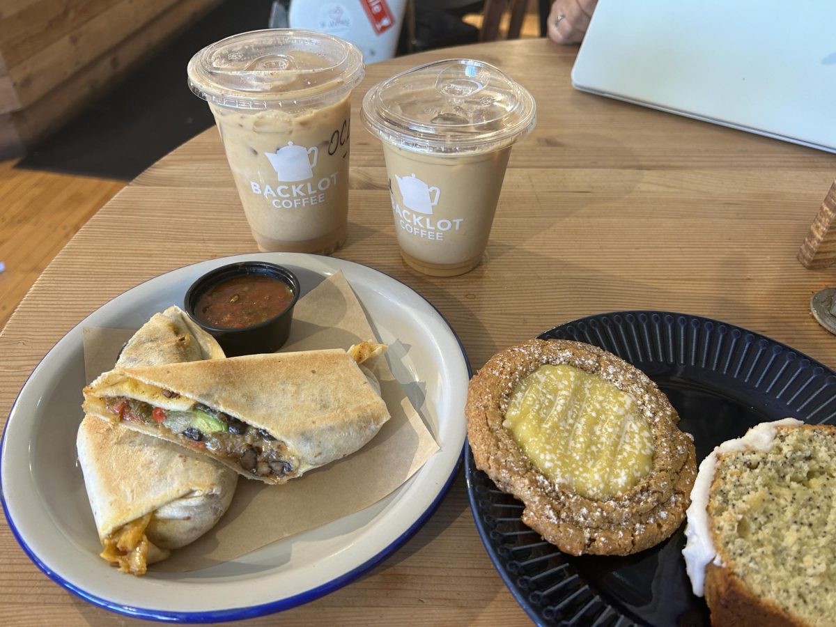 A table with an iced oatmeal cookie latte, an iced cardamom rose latte, a white plate with a Backlot veggie burrito and a navy plate with a key lime pie cookie and lemon poppyseed loaf.