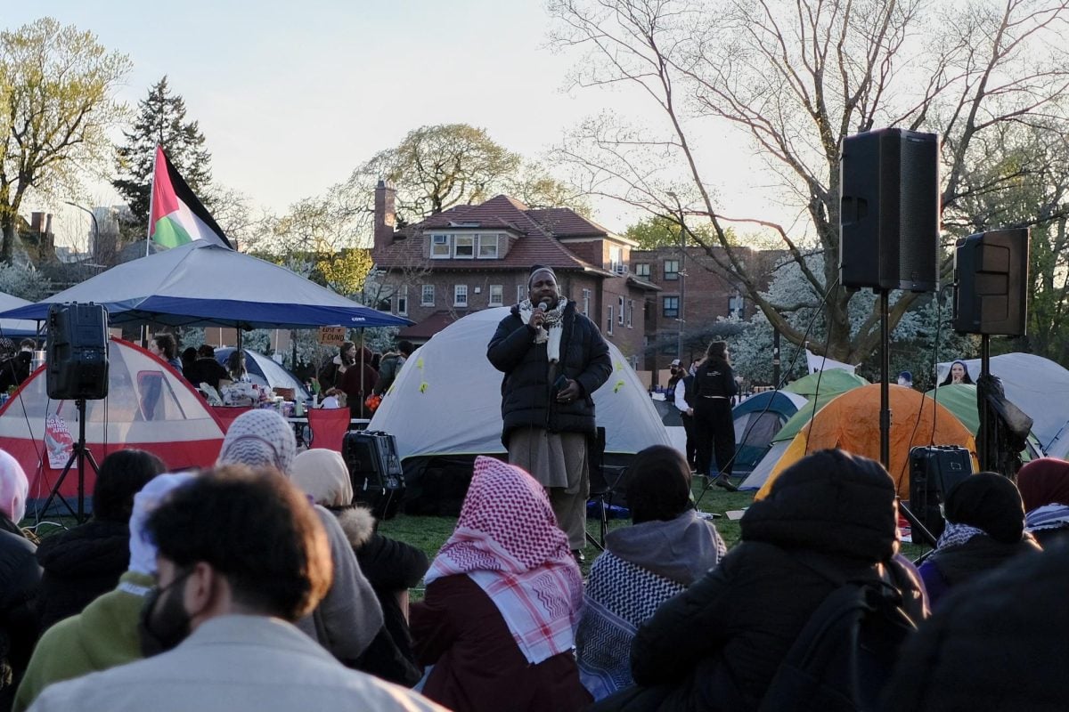 A man speaks, surrounded by a crowd and tents.