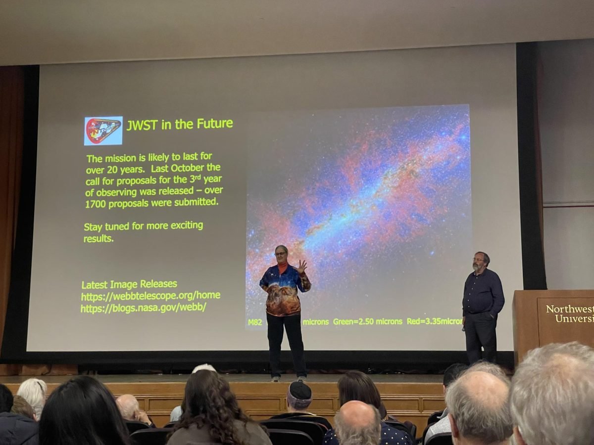 A woman stands in the center of the stage talking to an audience in front of a projection of a galaxy.