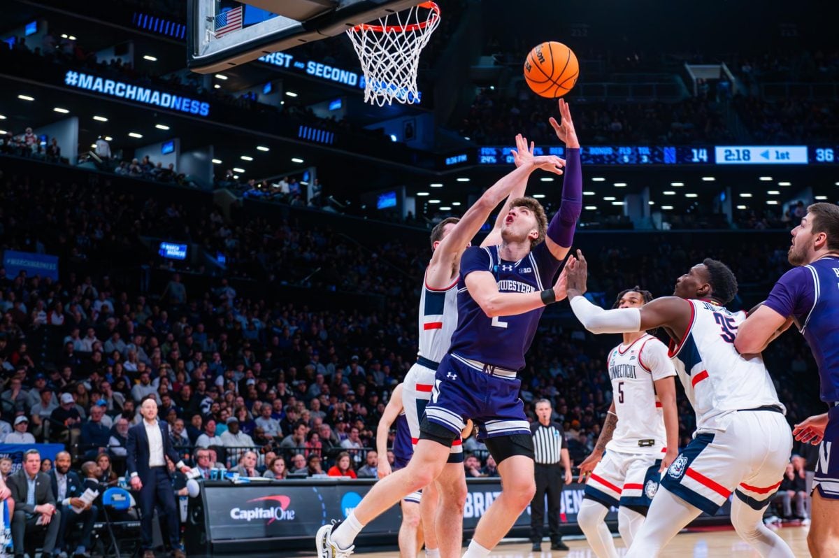 A basketball player in purple reaches out with their left arm to throw the ball into the basket.