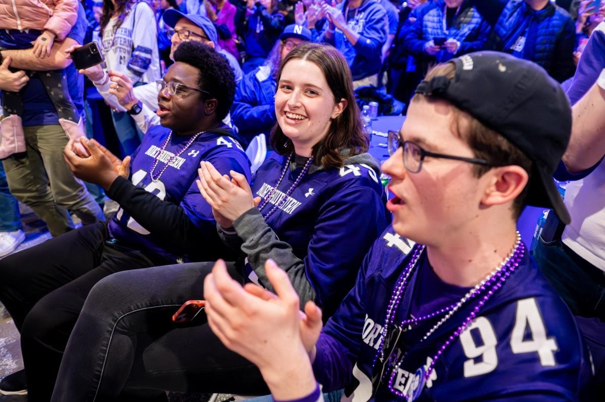 An audience member in a purple jersey looks at the camera while clapping.