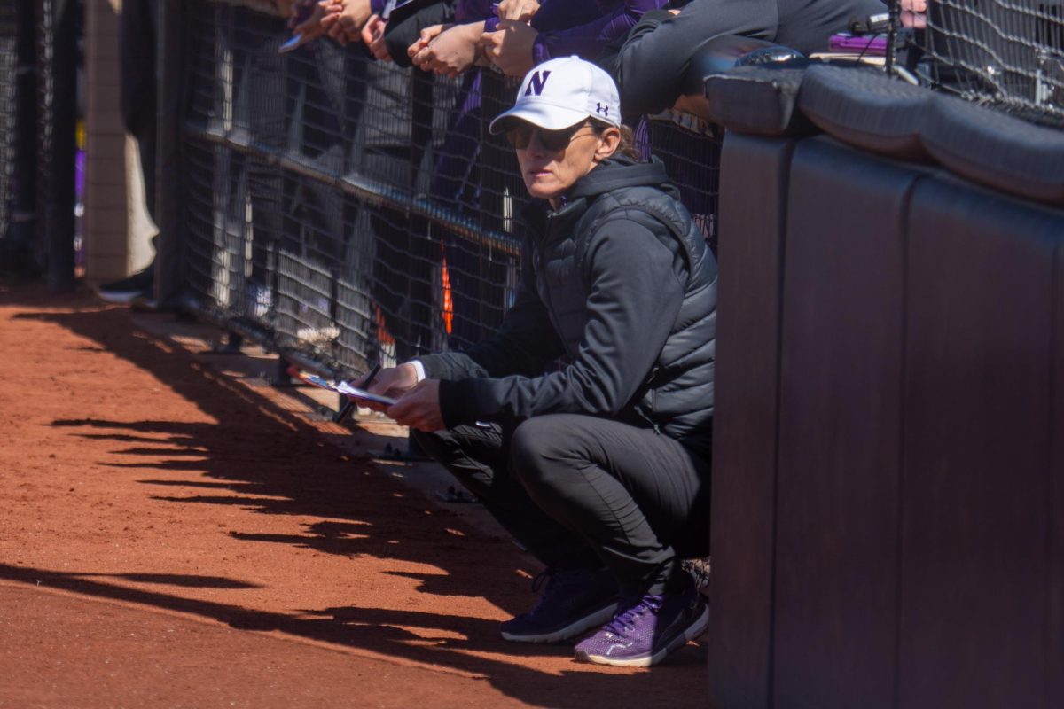Northwestern coach Kate Drohan squats by the dugout as she watches on-field action.