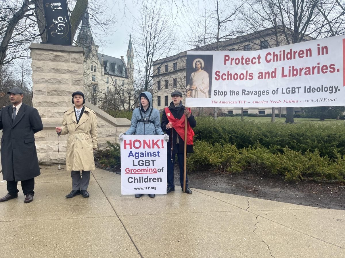 Four protesters stand beside Weber Arch with signs reading “HONK! Against LGBT Grooming of Children” and “Protect Children in Schools and Libraries.”