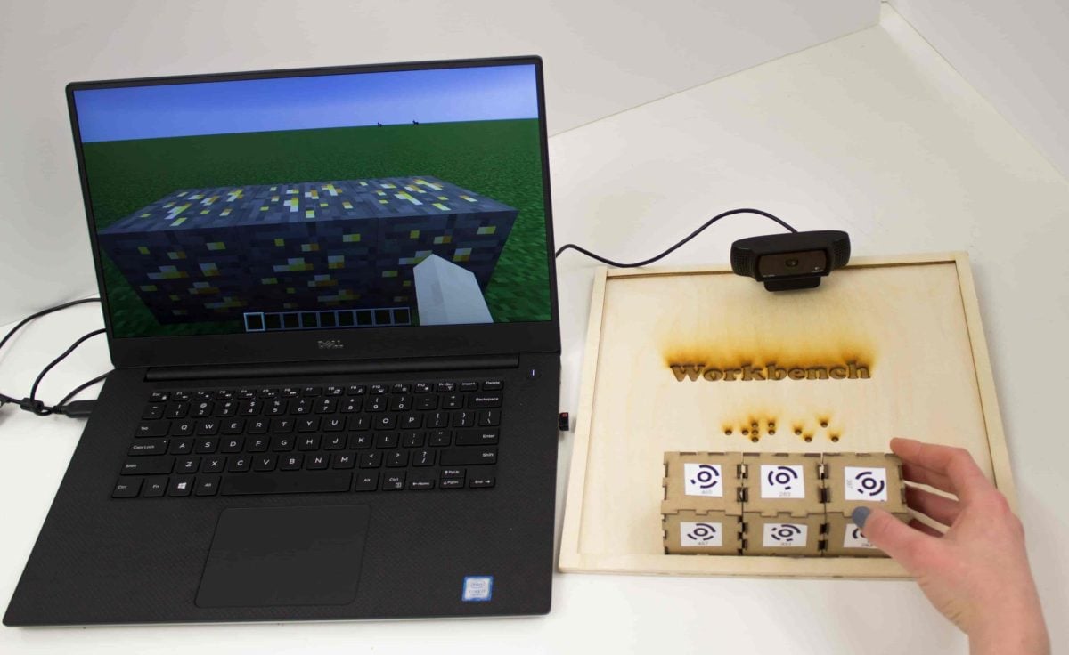 A computer with Minecraft on the screen lies next to a workbench with three wooden blocks placed horizontally on it.