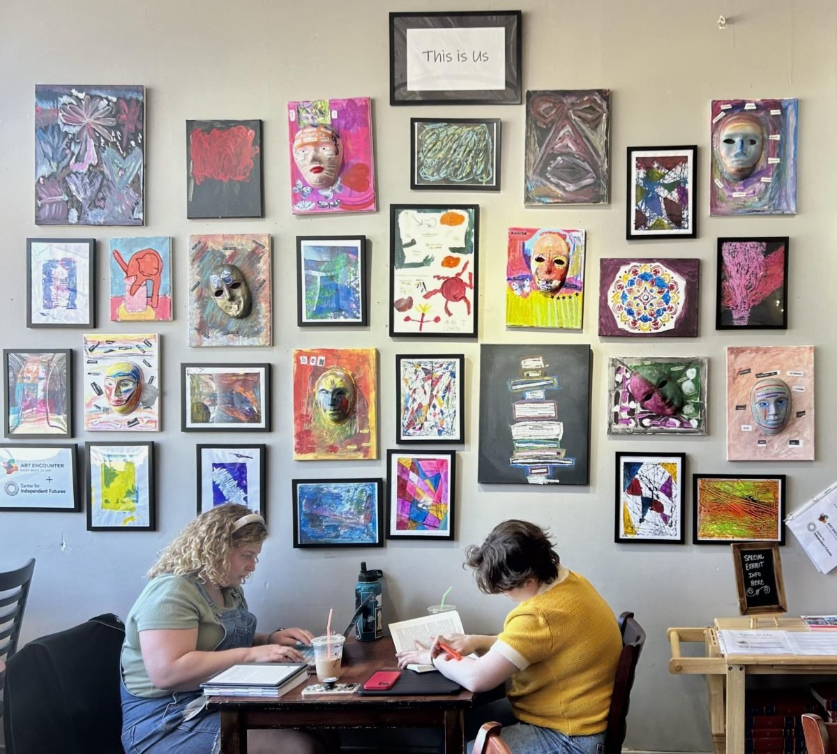 Two people sit at a table in front of a wall with several pieces of art on it.
