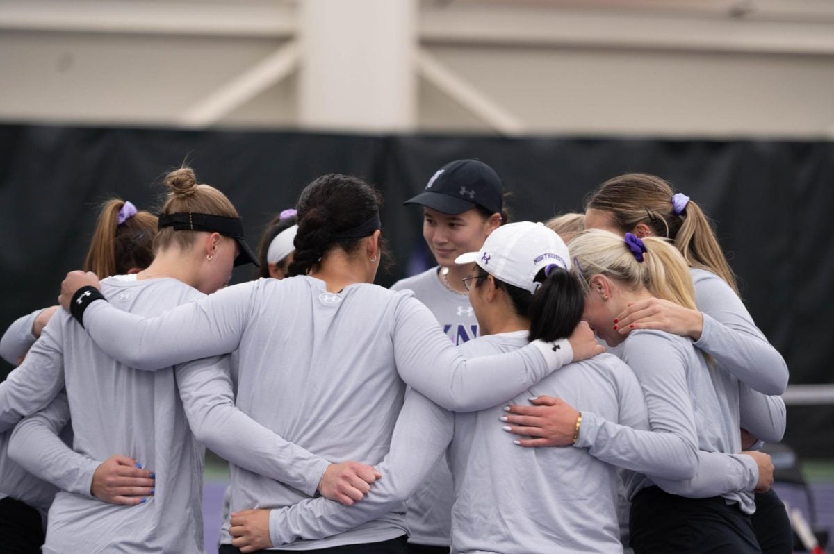 A group of tennis players wearing white/gray long-sleeved shirts gather together in a circle, their hands on each other’s shoulders.