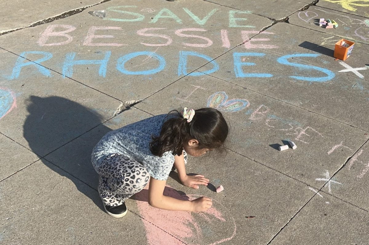 Student filling in a heart drawn on a sidewalk with pink chalk in front of messages reading “SAVE BESSIE RHODES” and “I (heart) BESSIE RHODES.”