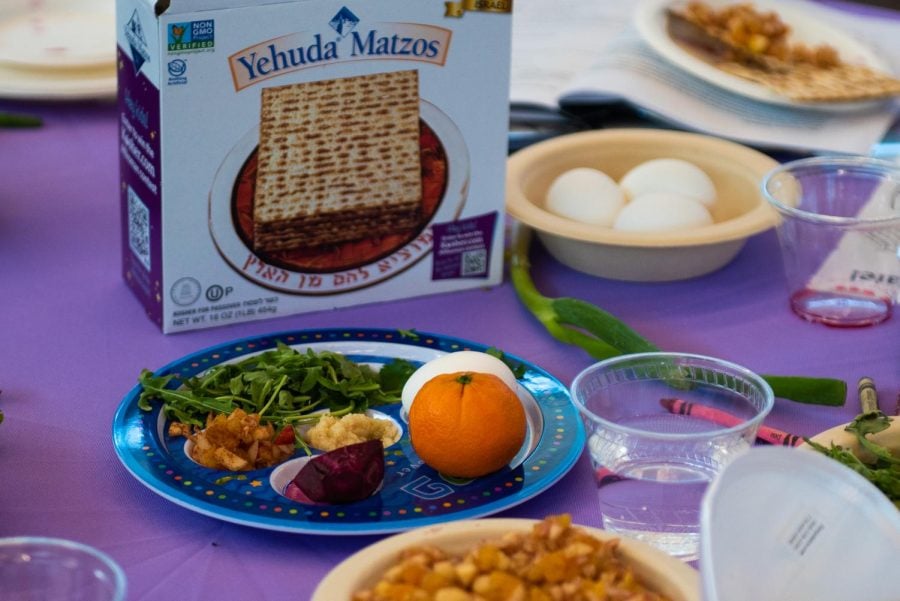 A seder plate sits in front of a matzo box, surrounded by water cups, other food and a crayon.