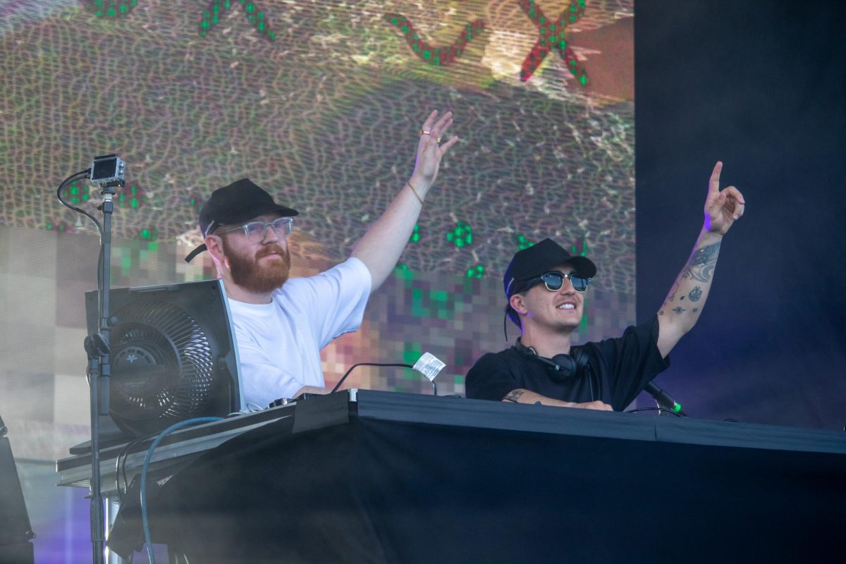 Two DJs onstage raise their left arms up while mixing behind a table. 