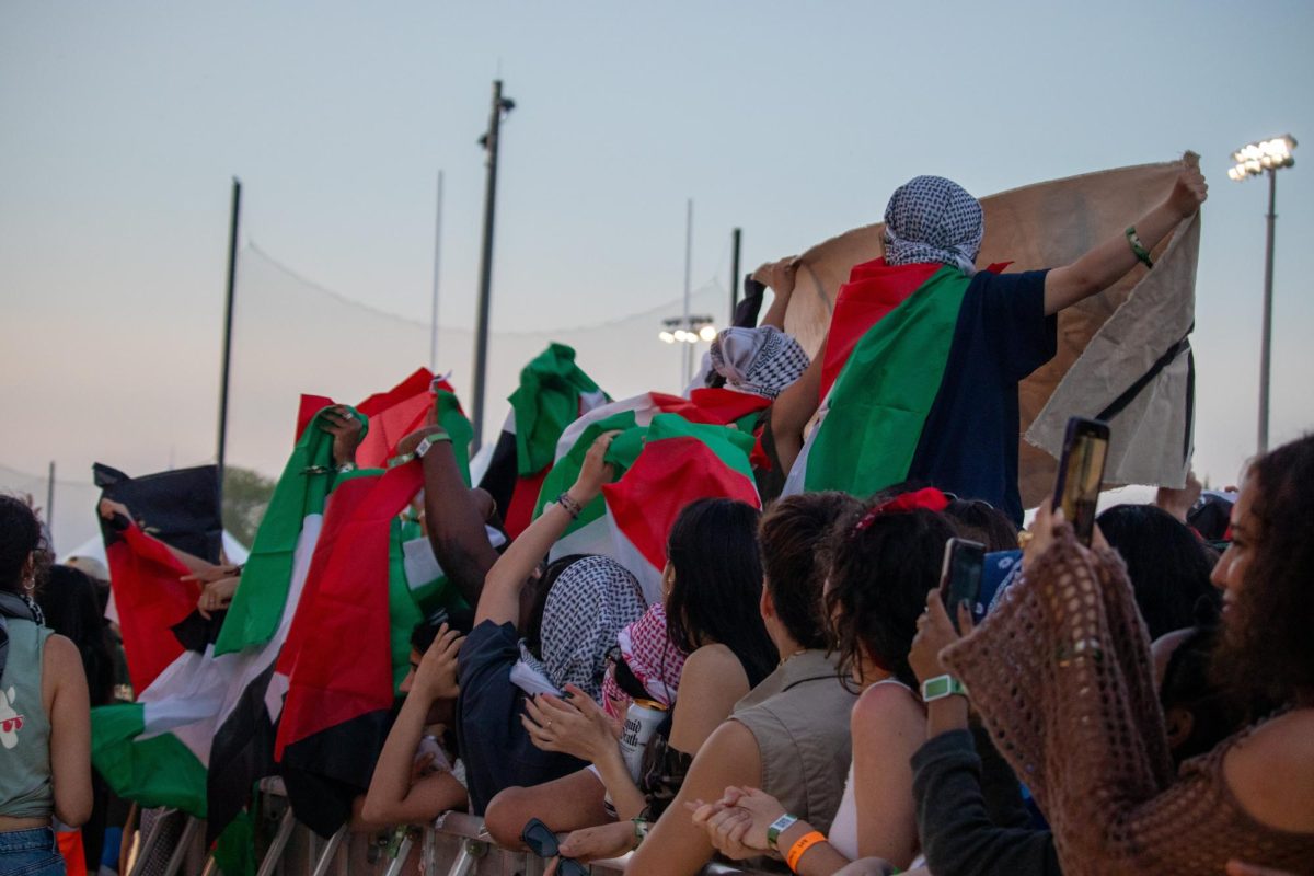 A group of people behind a concert barricade lift Palestinian flags. Some sit on the shoulders of others and hold up a banner while others on the side film with their cellphones.